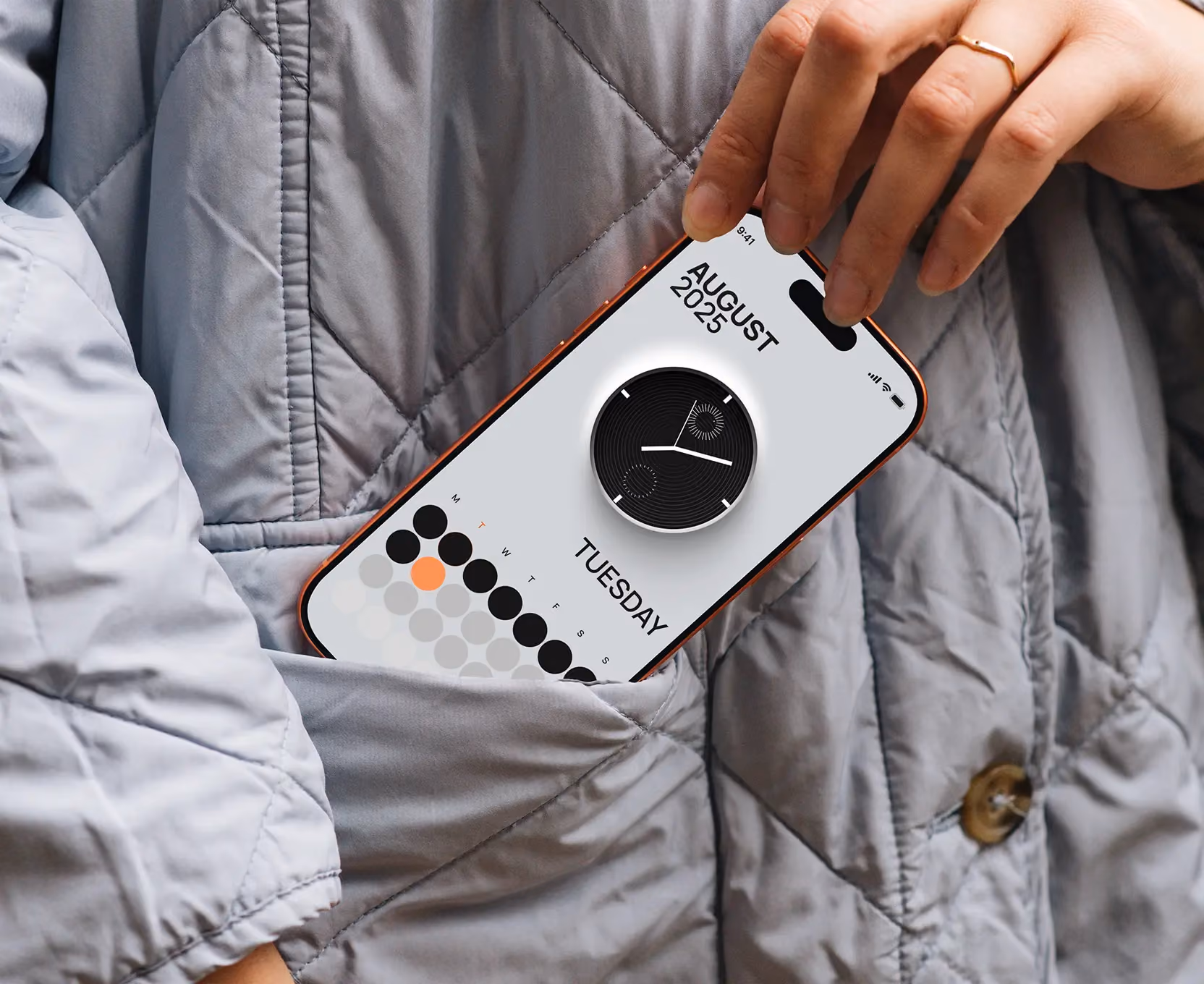 Hand placing a smartphone showing August 2025 calendar and a round clock into a gray quilted jacket pocket.