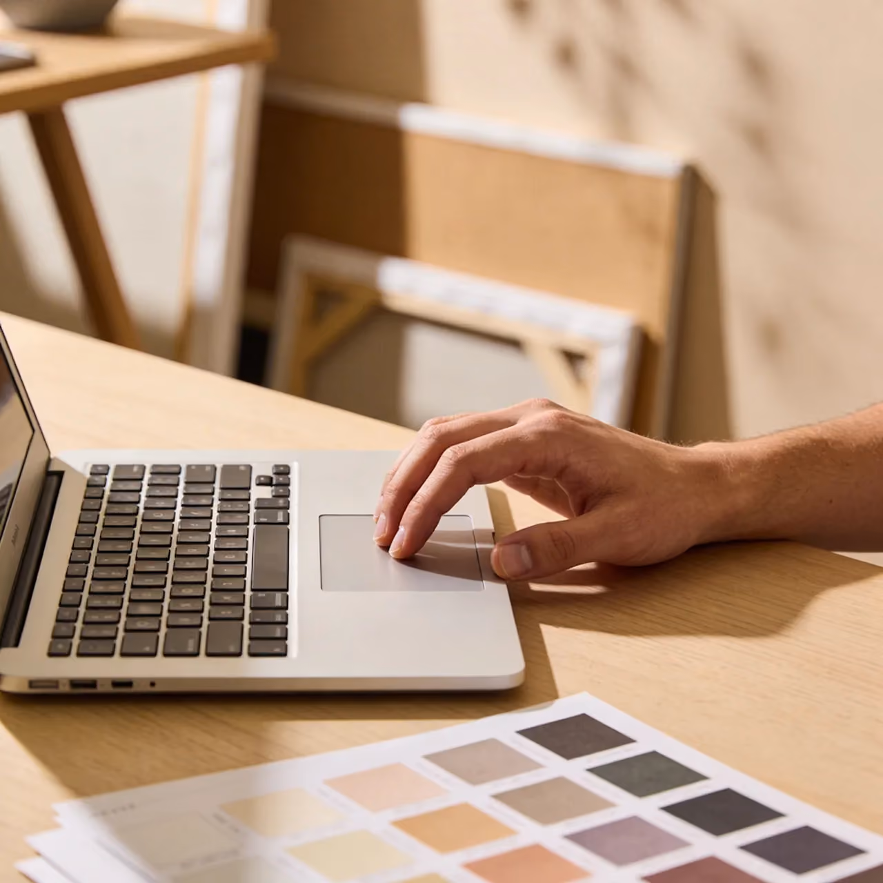 Hand using laptop trackpad next to color palette swatches on a wooden desk.