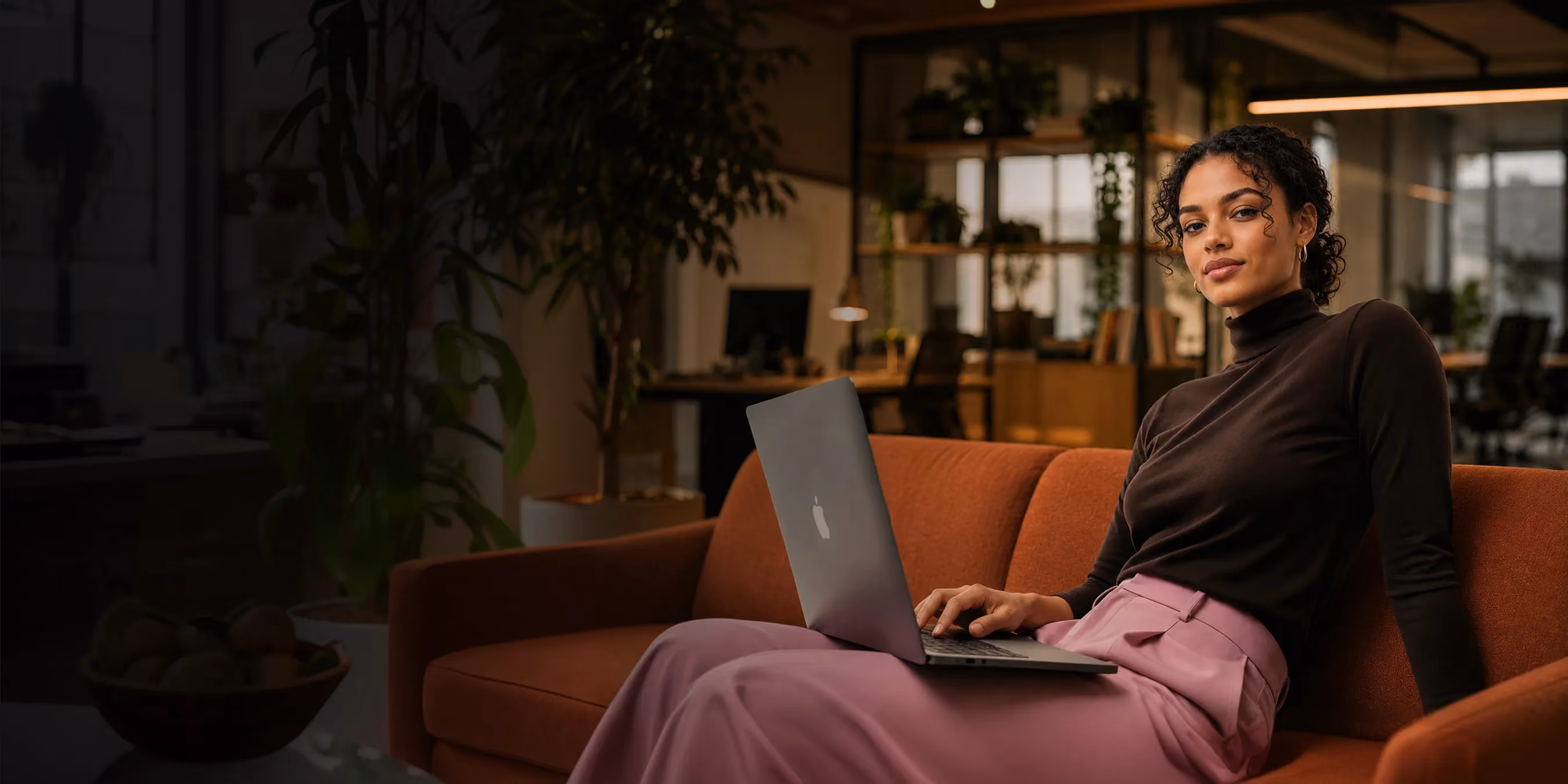 Woman with curly hair sitting on a brown couch using a laptop in a cozy office space.
