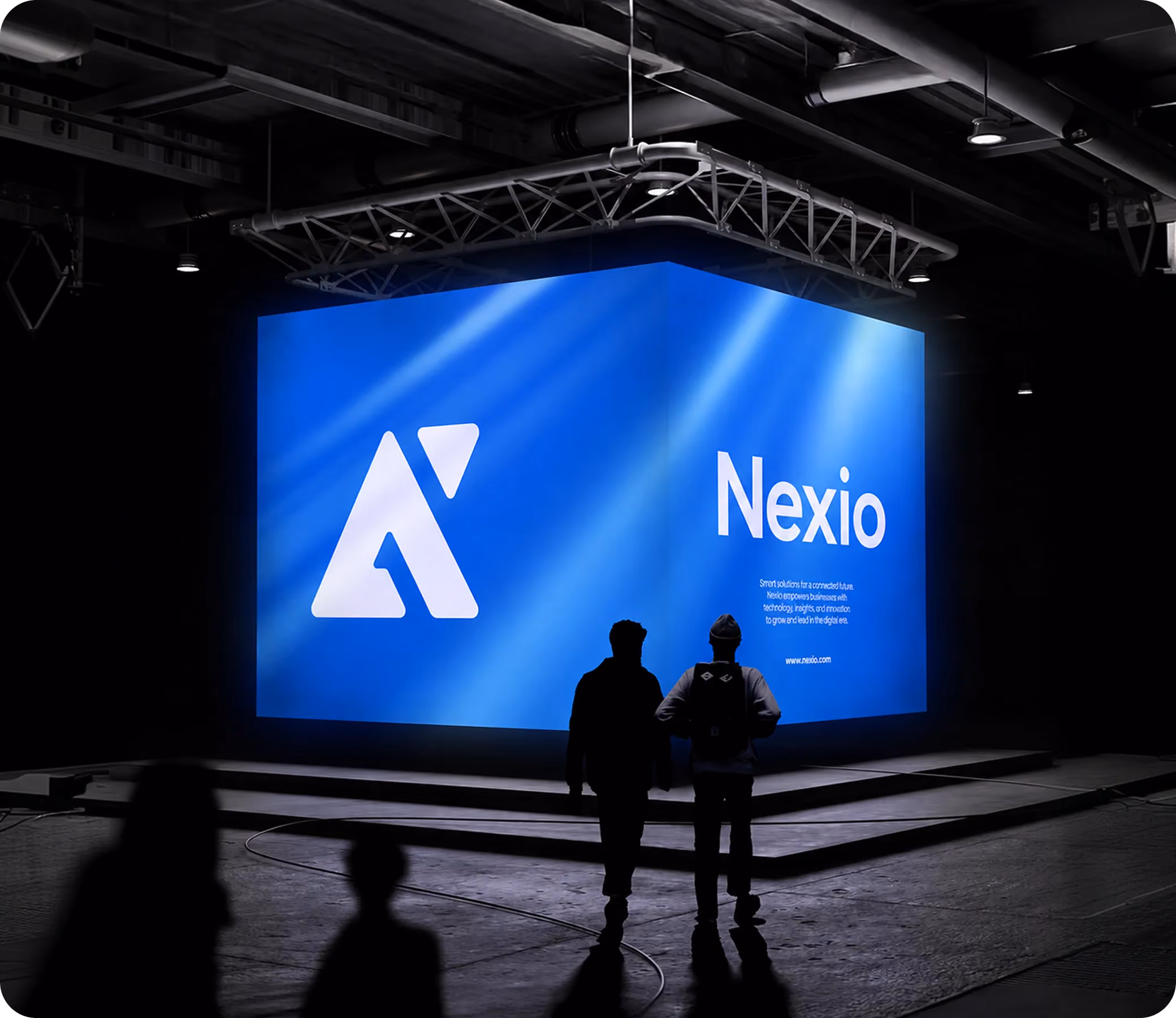 Two people stand in front of a large illuminated blue Nexio display in a dark indoor space.