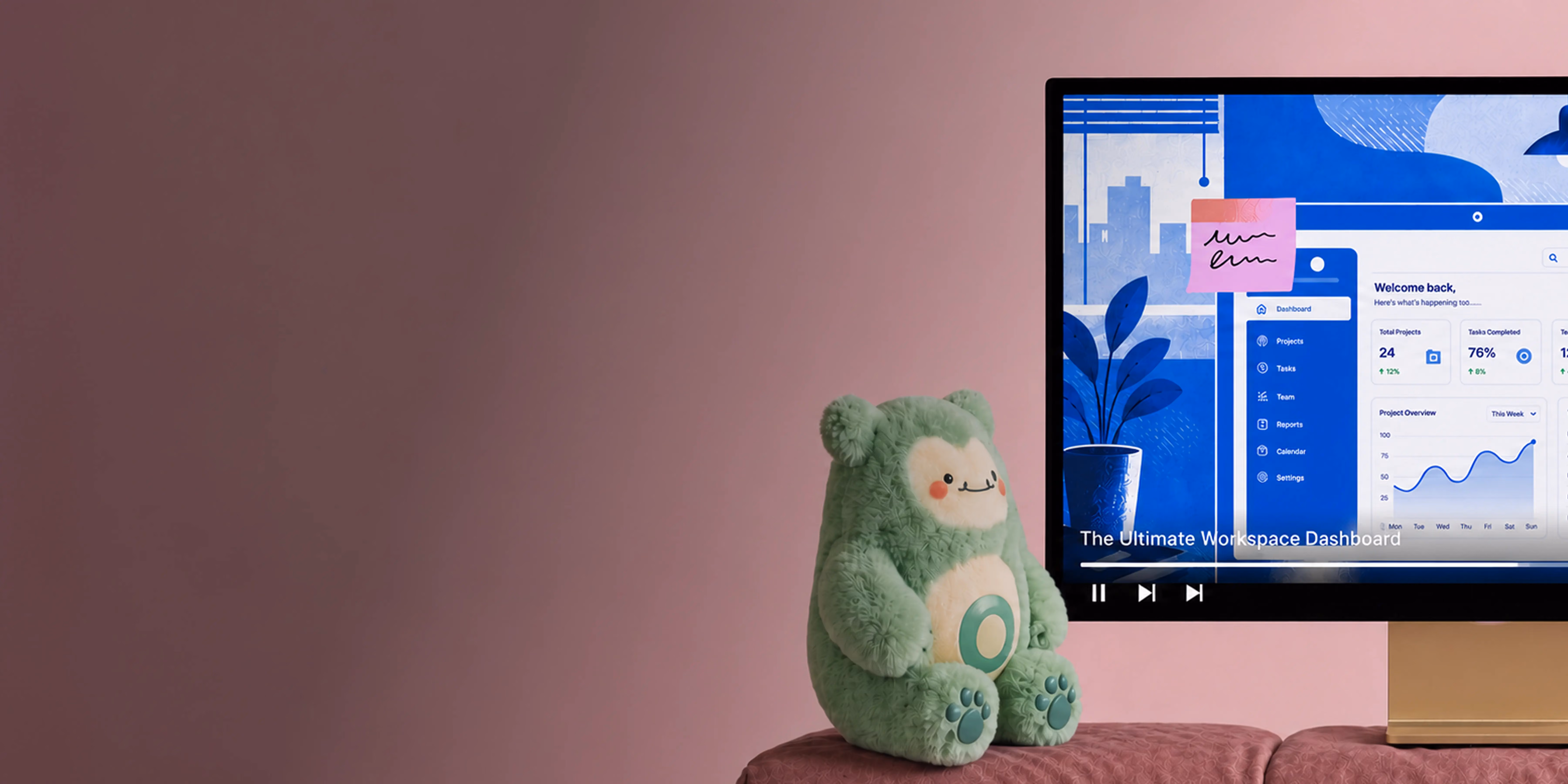 Green plush toy with a smiling face sitting on a pink cushion beside a TV screen showing a workspace dashboard.