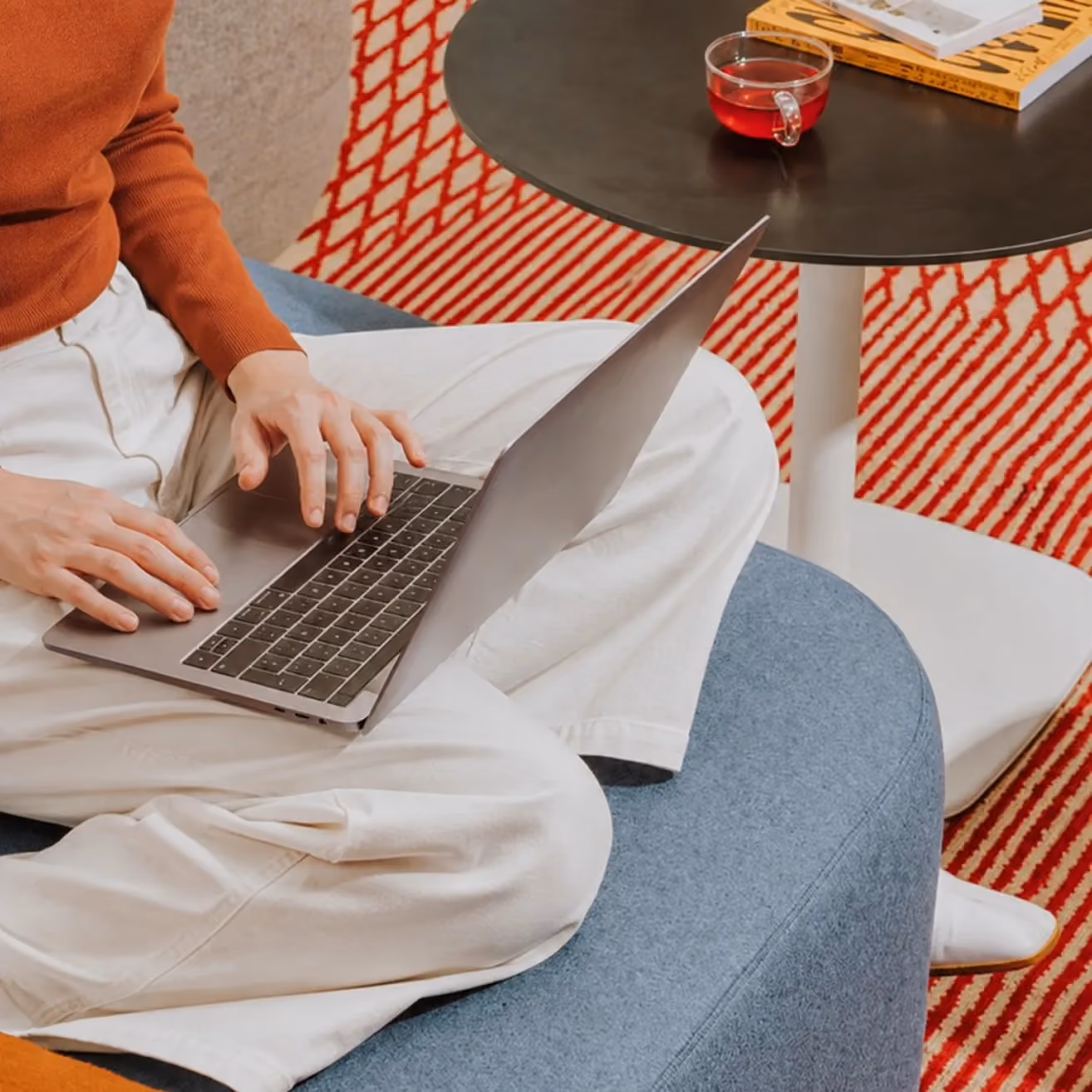 Person typing on a laptop while sitting cross-legged on a blue couch near a black round table with tea and books.