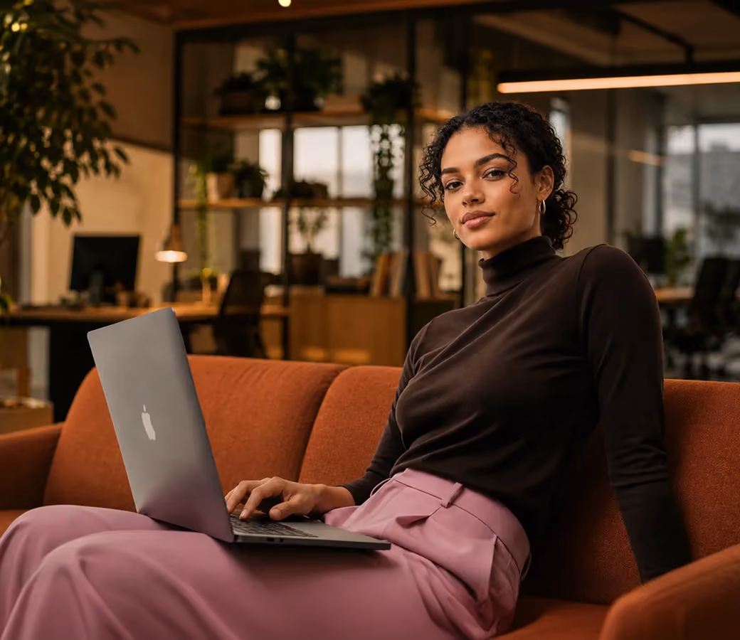 Woman in a black turtleneck and pink skirt working on a laptop while sitting on a brown couch in an office.