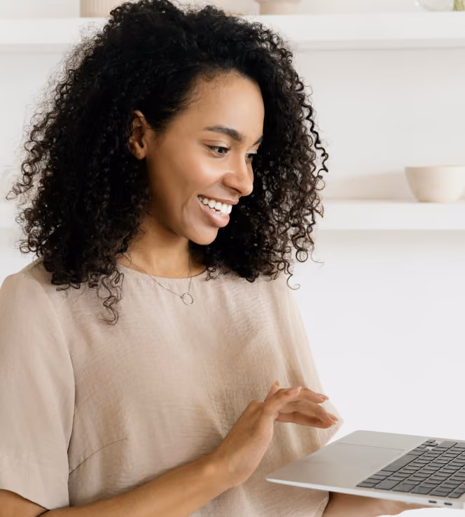 Smiling woman with curly hair using a laptop in a bright room with white shelves behind her.
