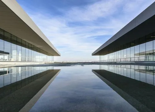Reflection of two modern glass buildings on calm water under a blue sky with clouds.