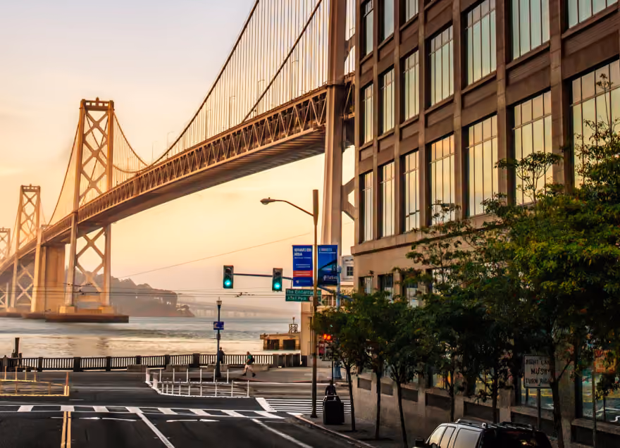 View of a large suspension bridge over water during sunset, with a city street and building in the foreground.