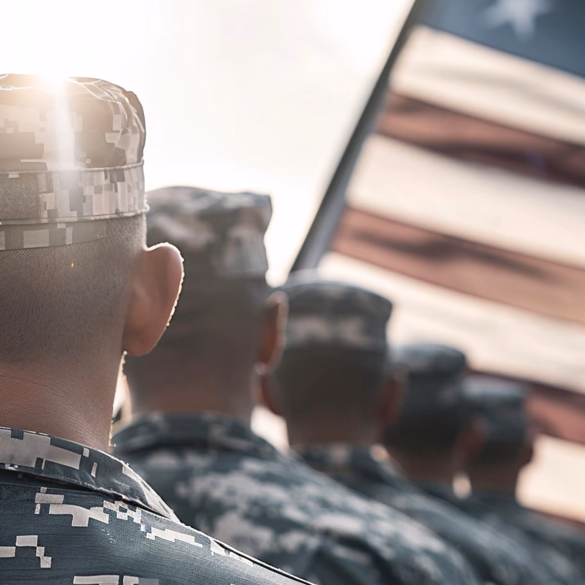 Rear view of soldiers in digital camouflage standing in line with a blurred American flag in the background.