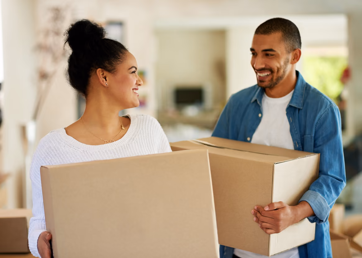 Smiling couple holding cardboard boxes, likely moving into a home together.