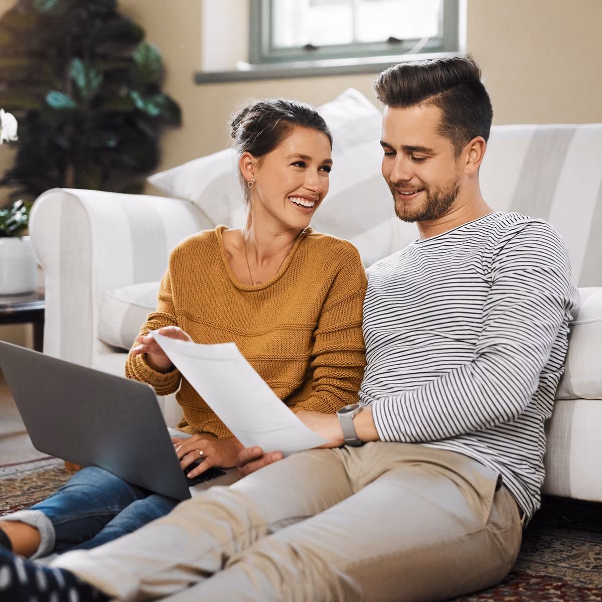 Smiling young couple sitting on floor by couch, reviewing documents and using a laptop together.