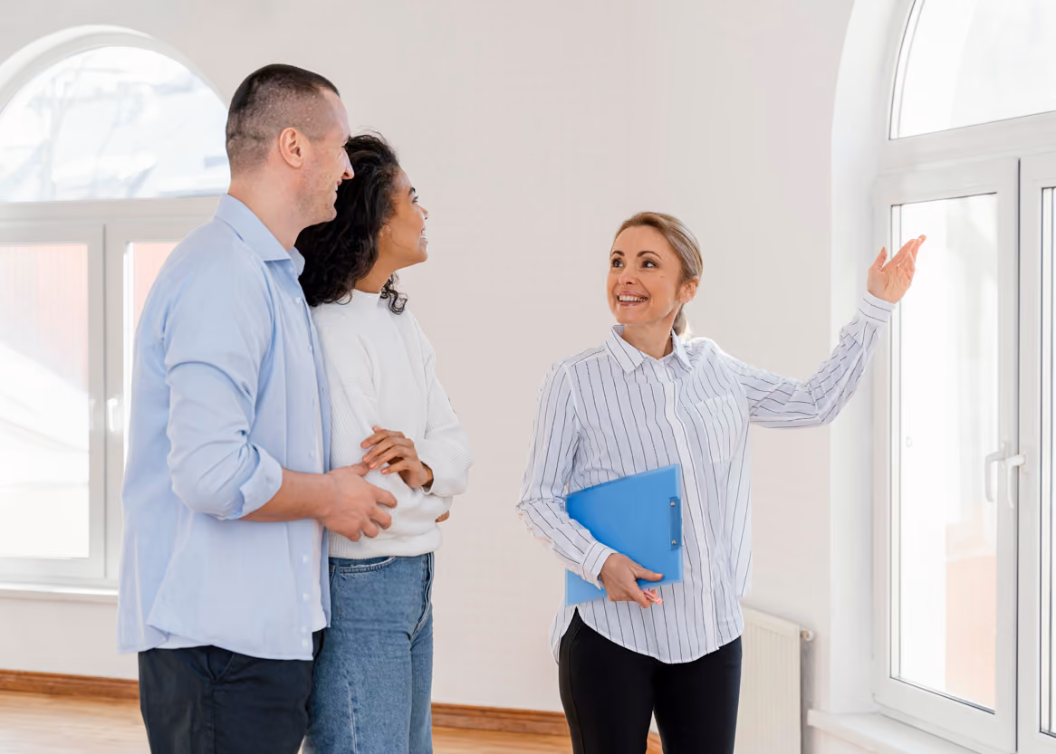 Female realtor smiling and showing an empty house to a young couple near a large window.