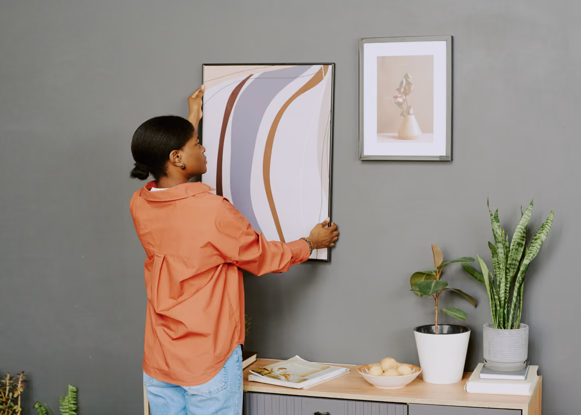 Woman in orange shirt hanging abstract art print with curved lines on gray wall.
