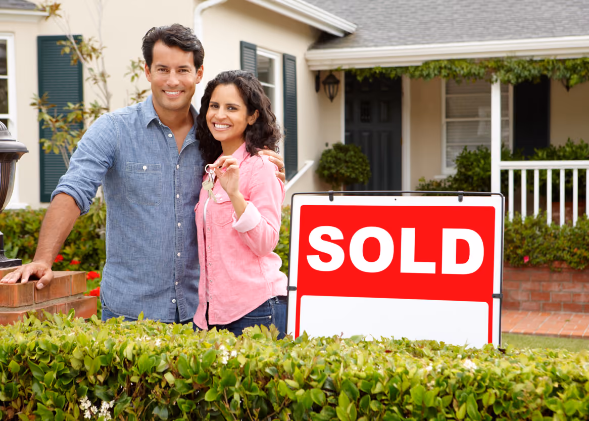 Smiling couple standing outside their home holding keys next to a large red and white sold sign.