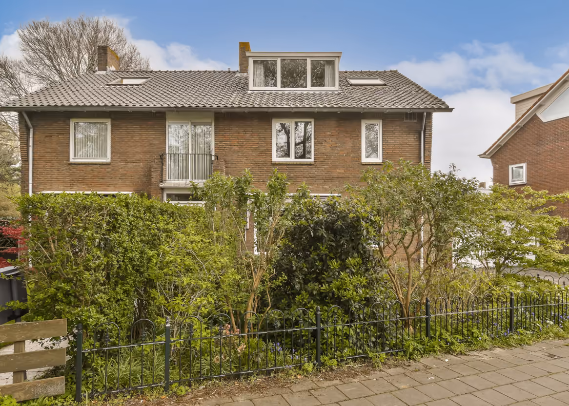 Two-story brick house with a tiled roof and green bushes in front behind a black metal fence.