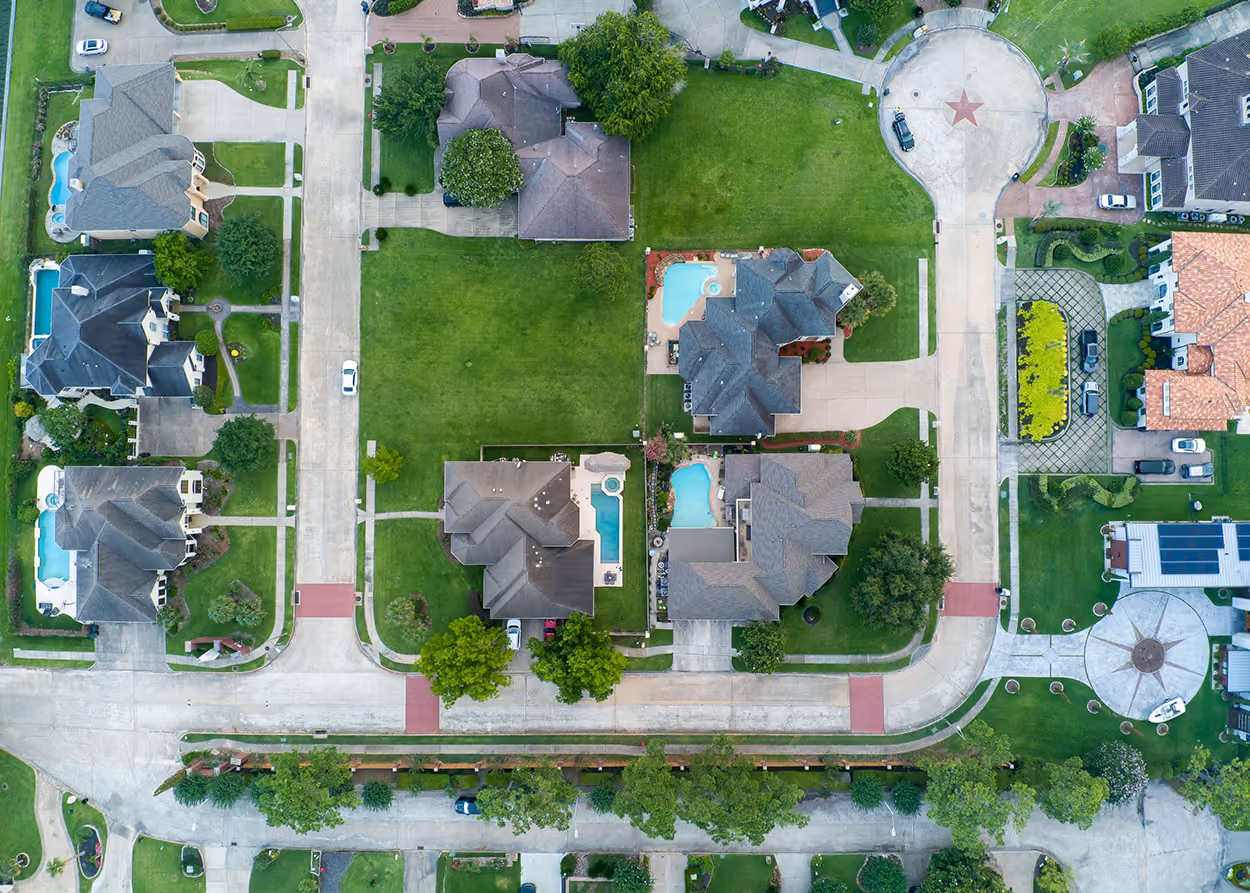 Aerial view of suburban neighborhood with houses, green lawns, swimming pools, and cars on clean streets.