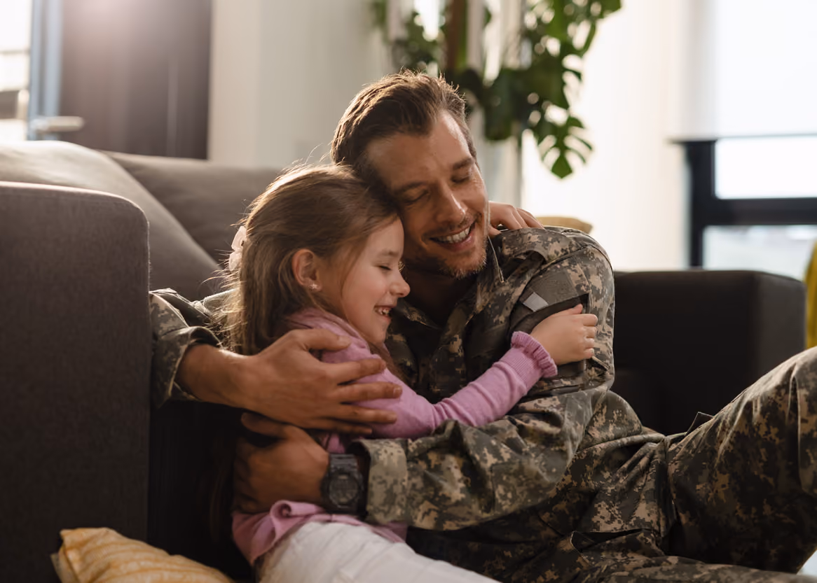 Smiling military father in uniform hugging his young daughter on the floor by a couch at home.