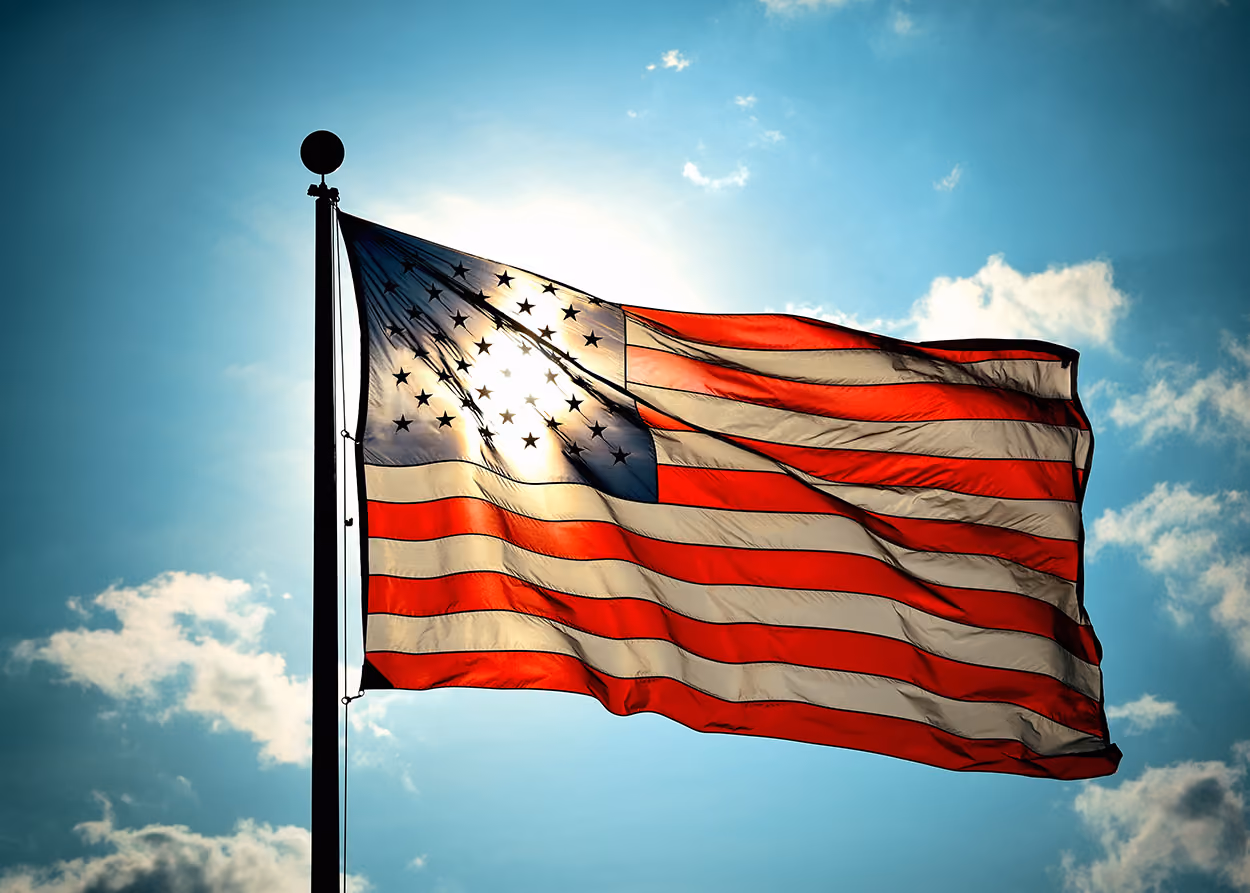 American flag waving on a pole with sun shining through it against a blue sky with clouds.
