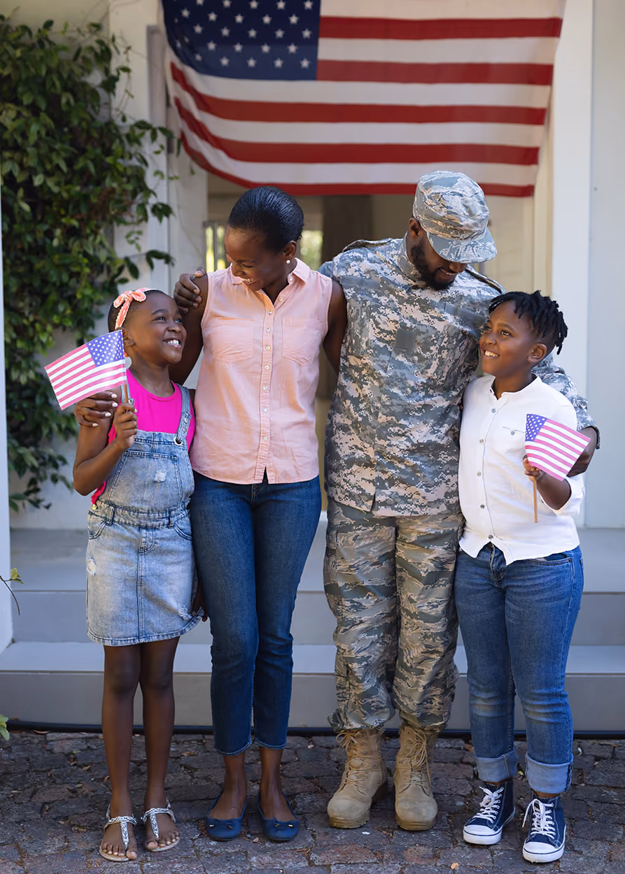 Smiling soldier embraces his family with mom and two kids holding American flags under a large flag.