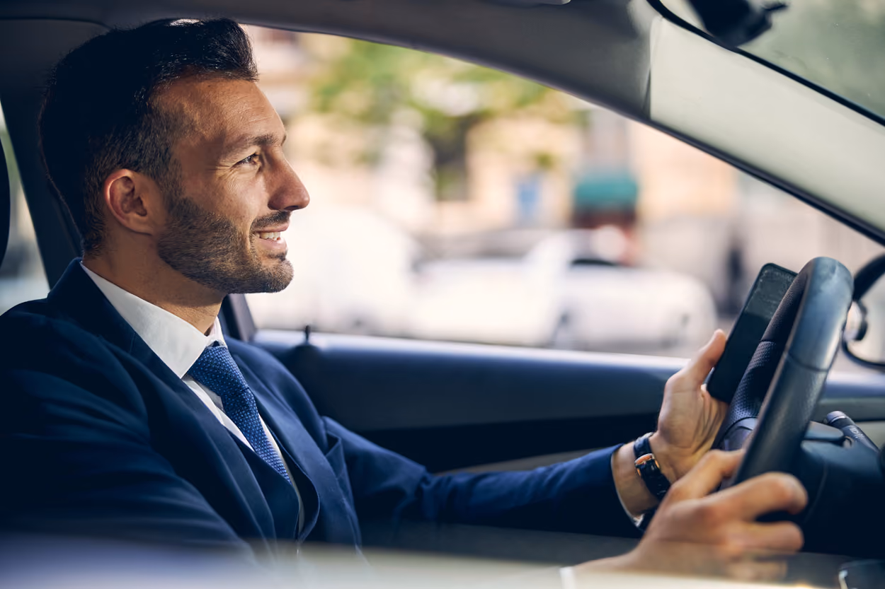 Smiling businessman in a suit driving a car while holding a mobile phone.