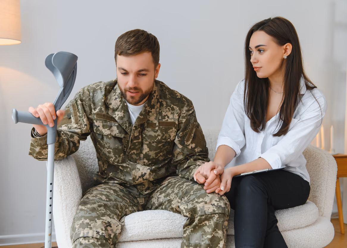 Sad soldier in camouflage holding crutches sits beside woman comforting him by holding his hand.