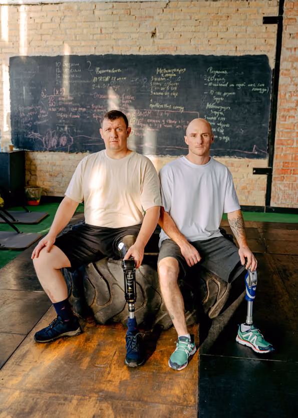 Two men with prosthetic legs sitting on a large tire in a gym with a chalkboard behind them.