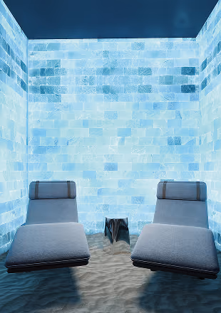 Two gray lounge chairs with headrests in a bright salt room with glowing salt brick walls and sand floor.