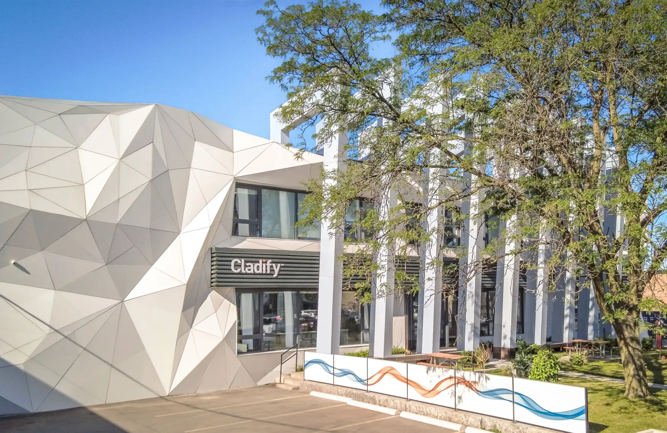 Modern office building with a geometric white facade on the left side and a series of tall white pillars in front of windows on the right side, partially obscured by a large tree with green leaves. The building has a sign that reads 'Cladify' above an entrance with stairs. In front of the building is a low barrier painted with flowing blue and orange wave designs, with an empty parking area in the foreground under a clear blue sky.