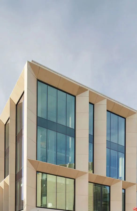 Modern building corner with large blue-tinted glass windows framed by beige concrete panels under a clear sky.