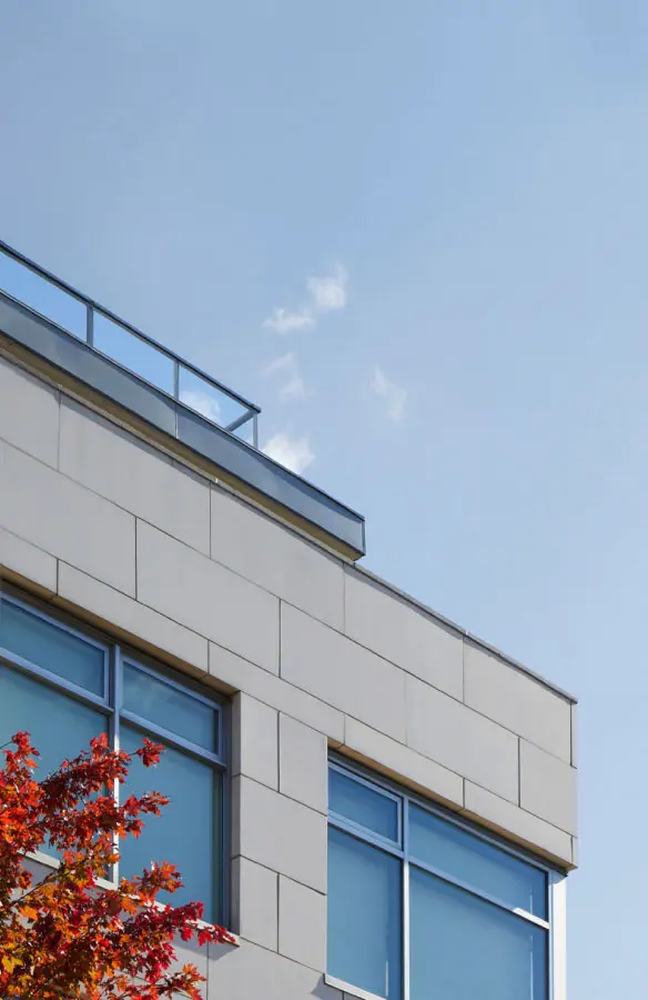 Corner of a modern building with large blue-tinted windows and light-colored stone cladding, with a tree with red leaves in the lower left and a clear blue sky in the background.