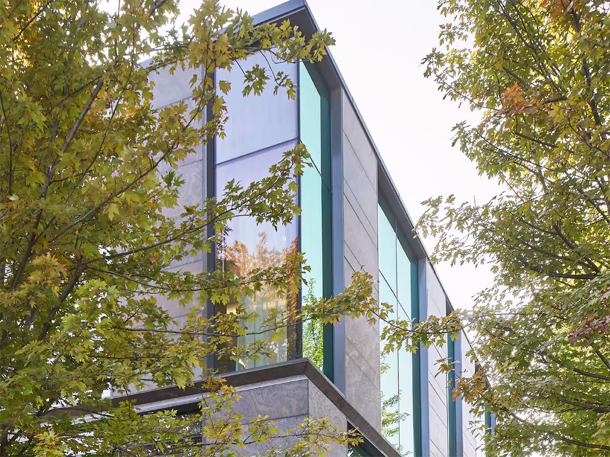 Modern building corner with large reflective glass windows framed by gray stone panels, surrounded by green leafy trees.