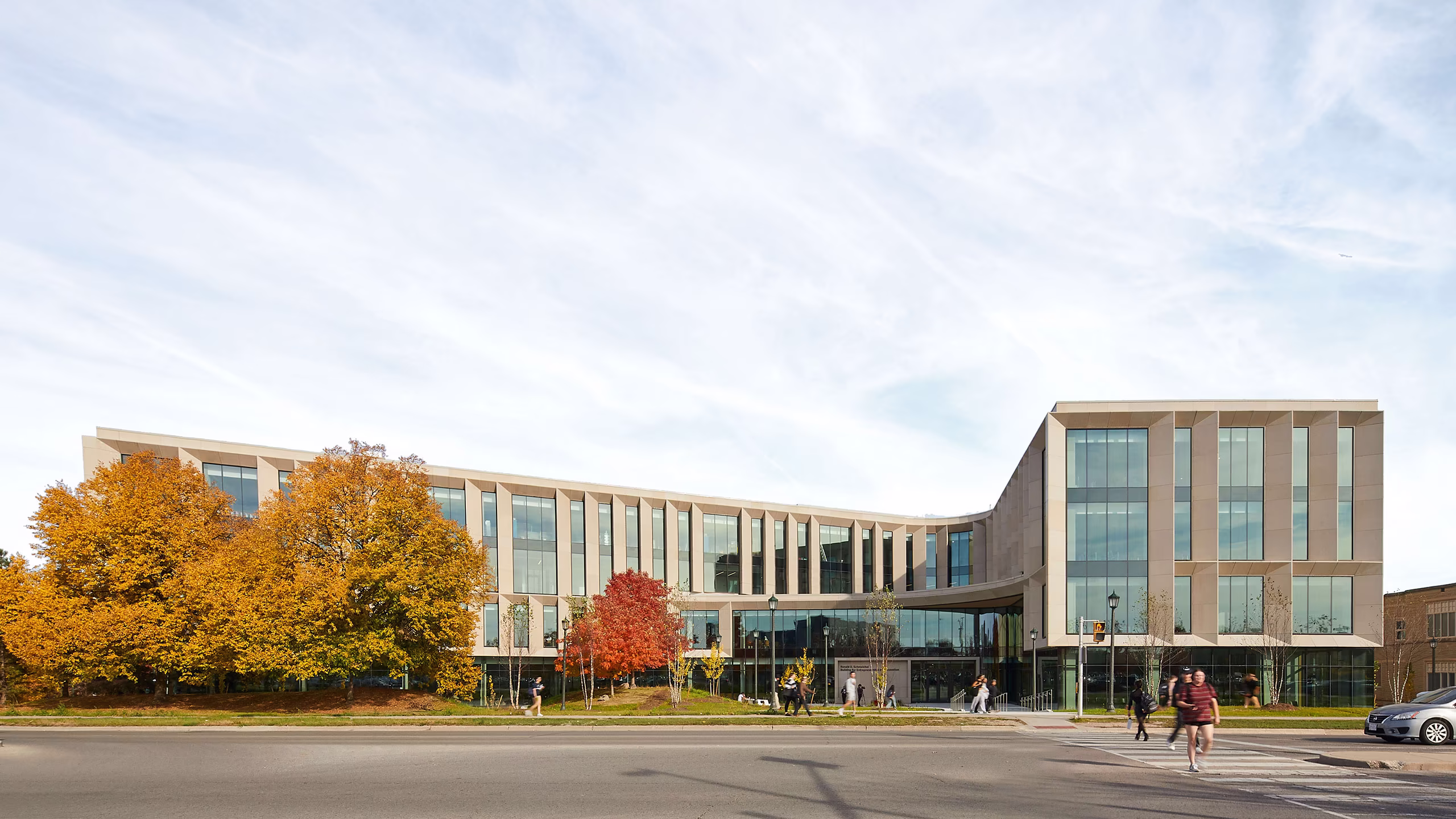 Modern three-story building with large vertical windows and light-colored exterior, surrounded by trees with autumn foliage, pedestrians crossing a street in front on a partly cloudy day.