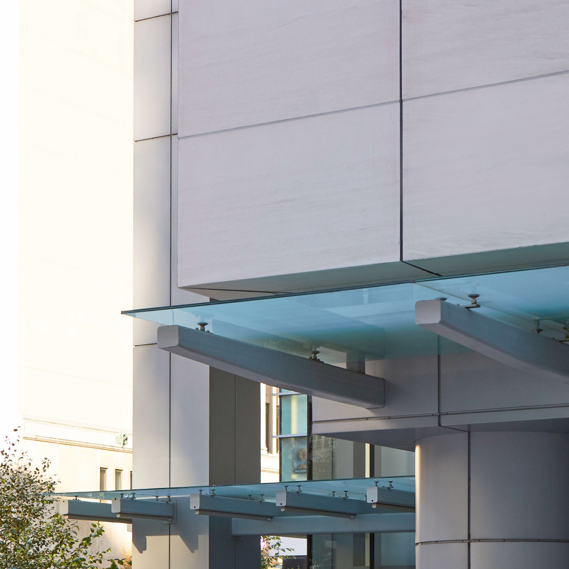 Modern building exterior featuring large glass awnings supported by metal beams attached to concrete walls.