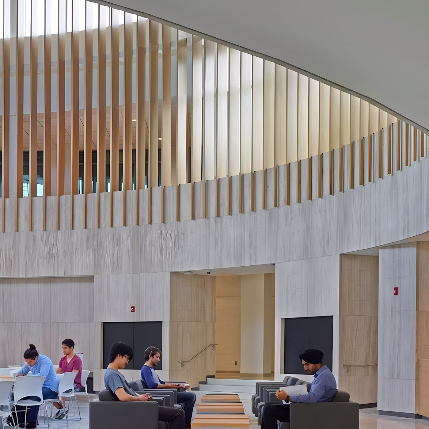Modern indoor space with a curved upper balcony featuring vertical wooden slats and several people sitting in chairs working or reading in a calm setting.
