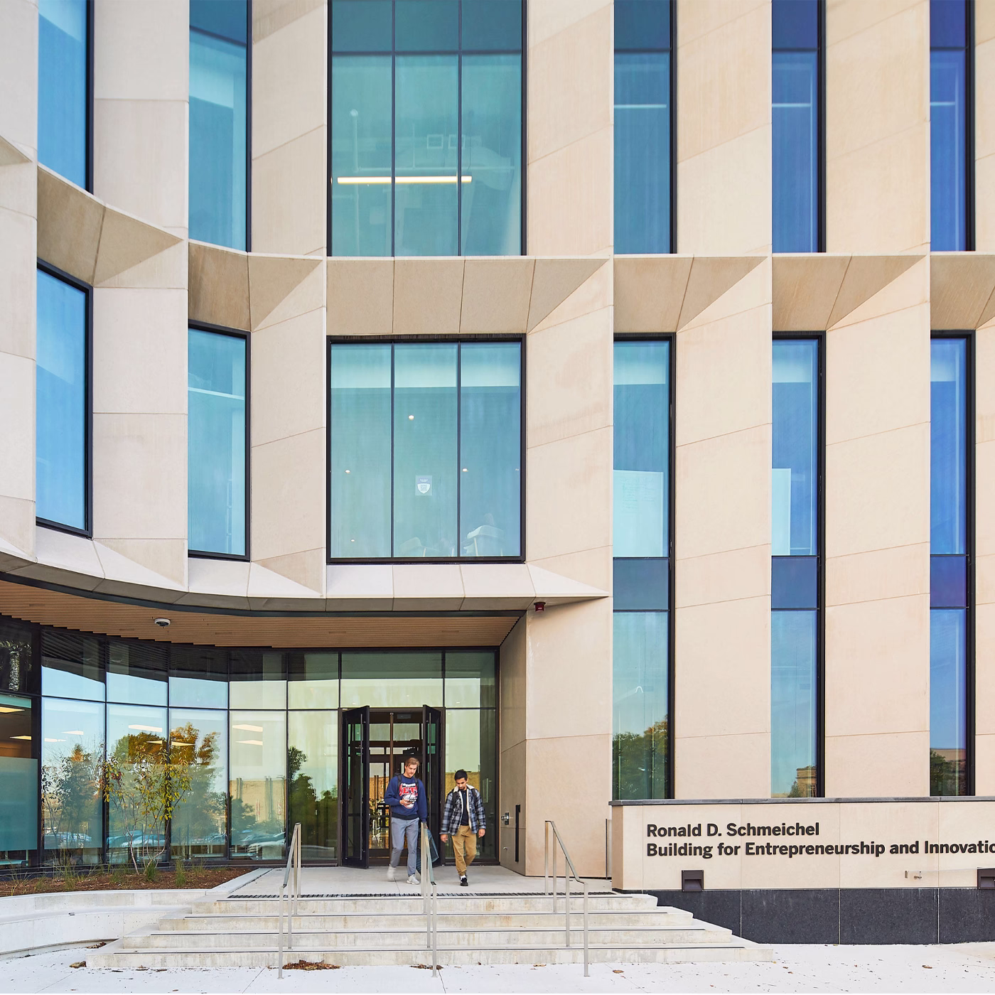 Modern building entrance with geometric light-colored facade and tall windows, showing two people walking down the steps. The building is named Ronald D. Schmeichel Building for Entrepreneurship and Innovation.