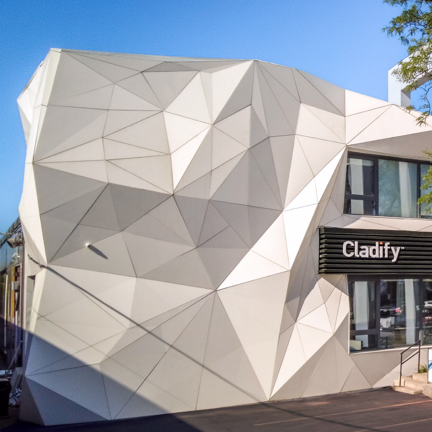Modern building facade with an angular, geometric design featuring interconnected triangular panels in shades of white and light gray under a clear blue sky, with a black sign that reads 'Cladify' on the right side above windows.