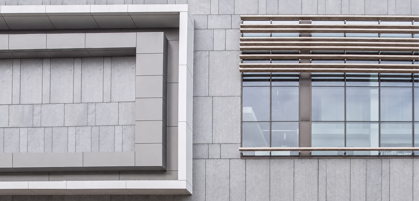 Close-up of a modern building facade showing gray rectangular stone tiles, a large window with horizontal wooden slats as sunshades, and a geometric frame feature on the left side.