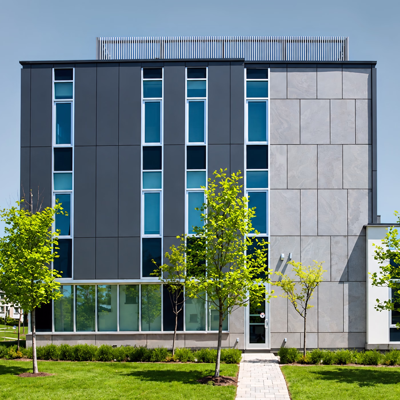 Modern three-story building with gray and stone facade, tall narrow windows, front door, and green lawn with small trees in front.