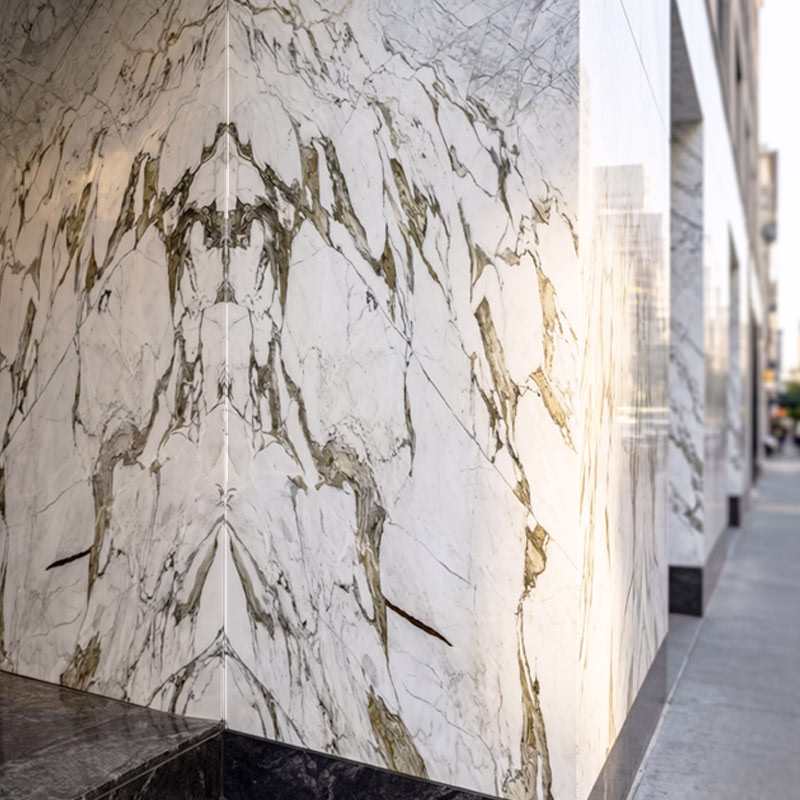 Close-up of a polished corner of a building exterior clad in white marble with grey and gold veining, with the sidewalk and street visible on the right.