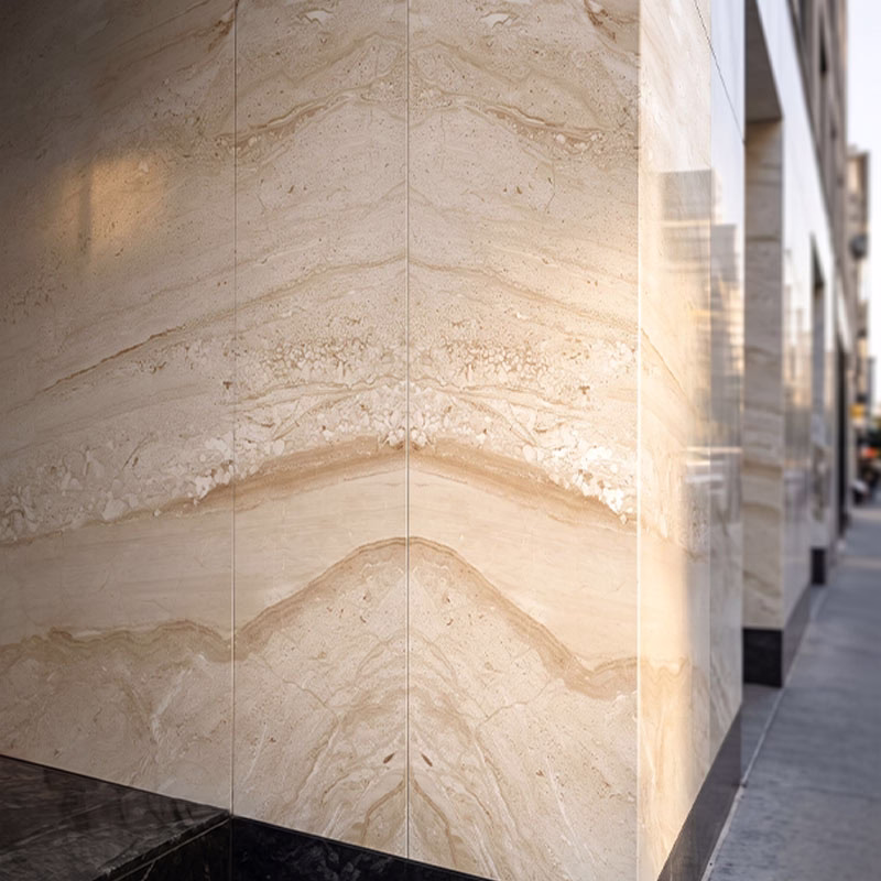 Close-up of a building exterior featuring polished beige stone tiles with natural veining, meeting at a corner above a black base, with a blurred urban sidewalk scene on the right.