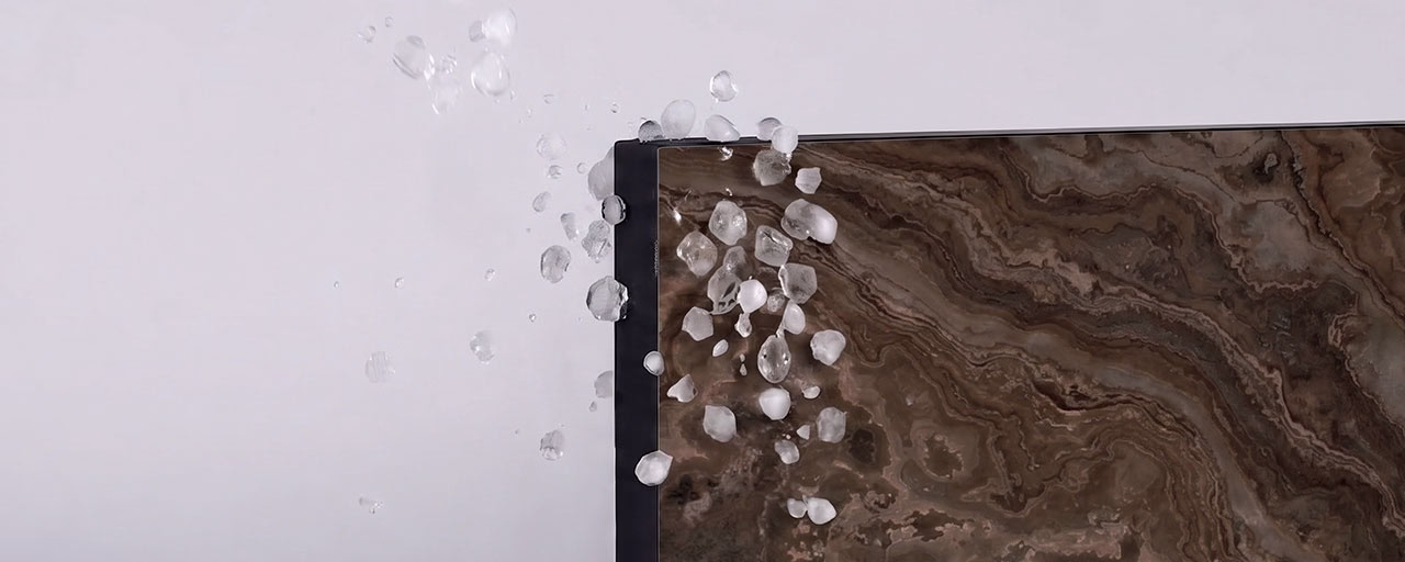 Close-up of ice pellets or hailstones bouncing off the edge of a smooth, glossy surface with a brown marbled pattern and black border against a plain light background.