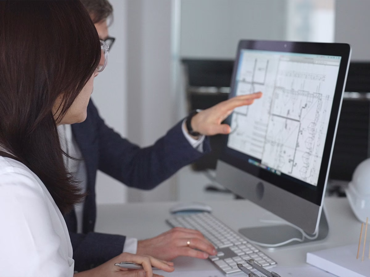 Two people working together at a desk, looking at architectural plans displayed on a computer screen with one person pointing at the screen.