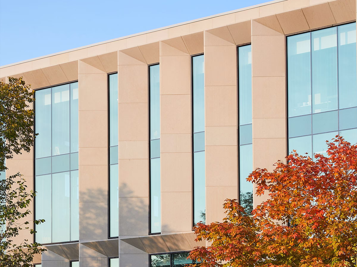 Modern building facade with large vertical glass windows separated by beige stone columns, with trees on either side, one showing autumn foliage.