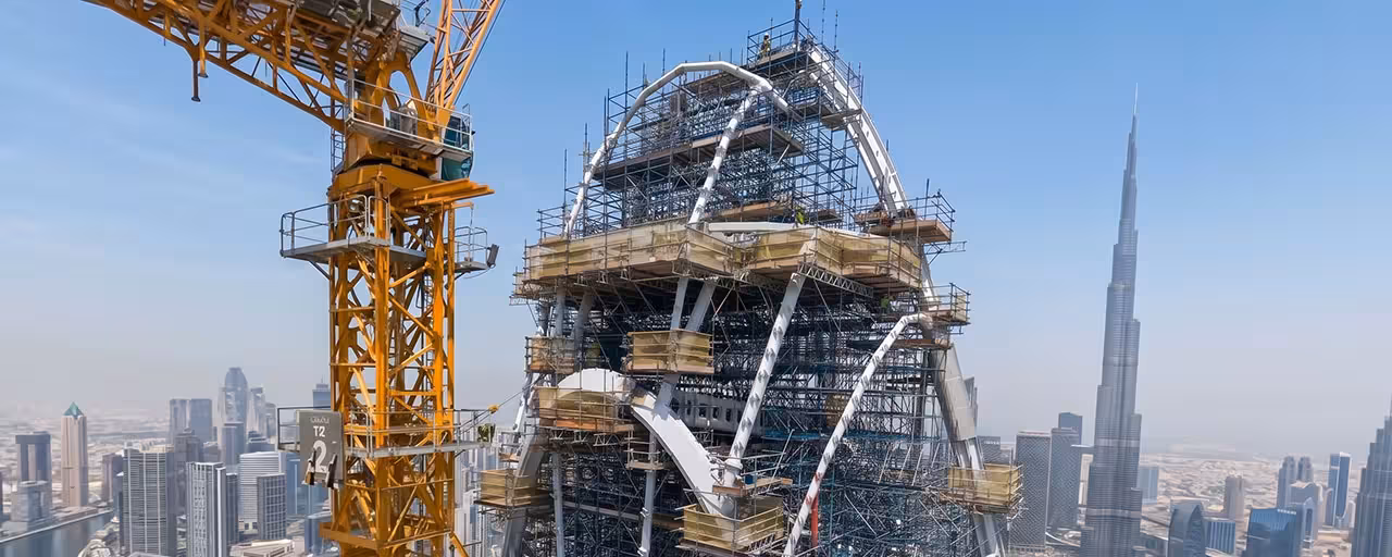 Construction site of a high-rise building with complex scaffolding and curved steel beams, a large yellow crane on the left, and a city skyline including the Burj Khalifa in the background under a clear blue sky.