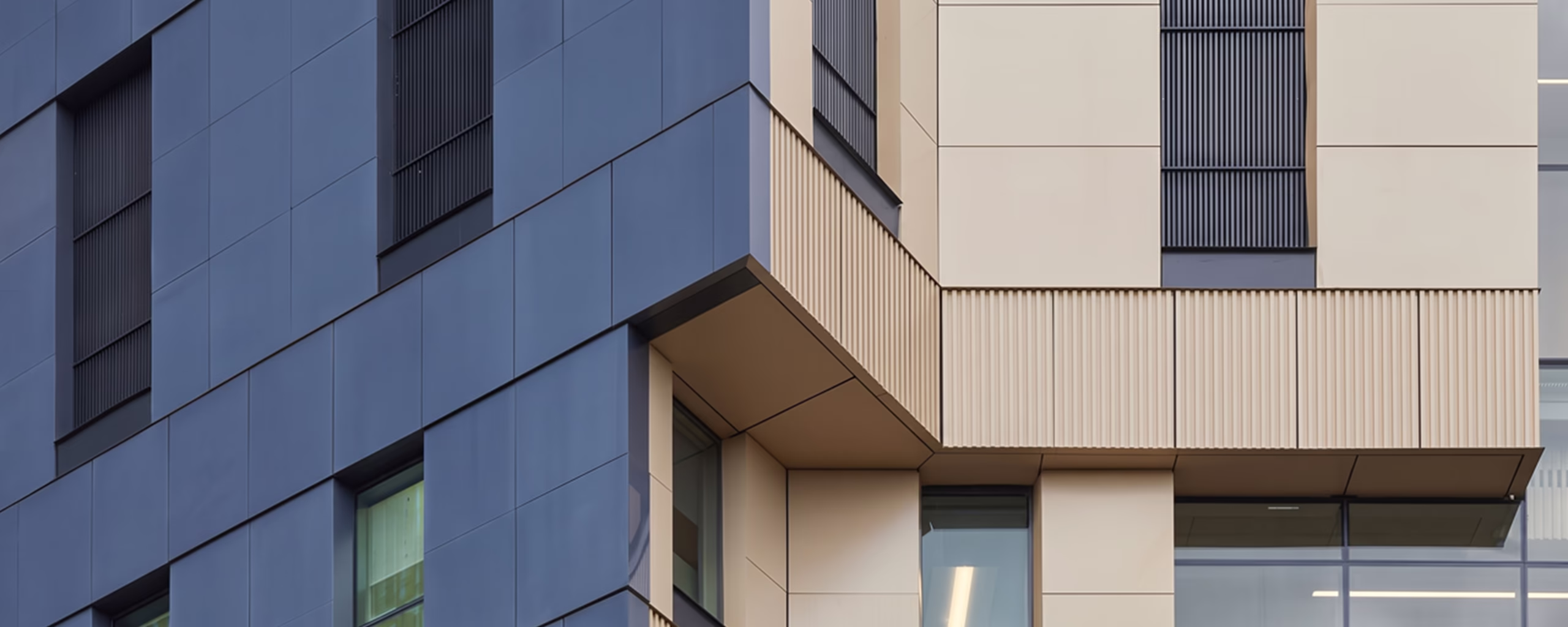 Close-up view of a modern building's exterior corner featuring dark blue and beige paneling with windows and vertical ventilation grilles.