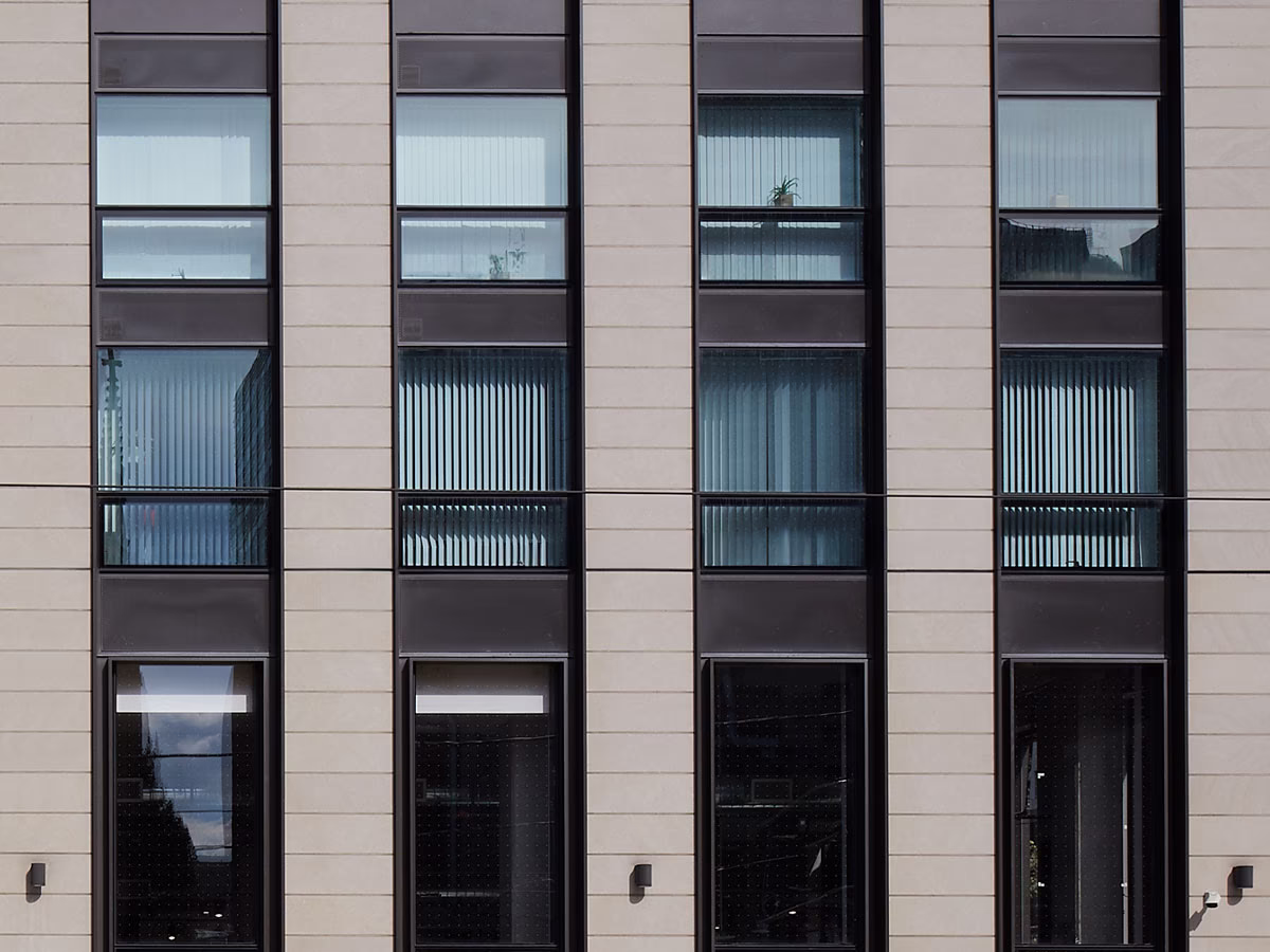Facade of a modern building with four vertical rows of tall windows framed by beige concrete and dark brown panels, some windows showing reflection and vertical blinds inside.