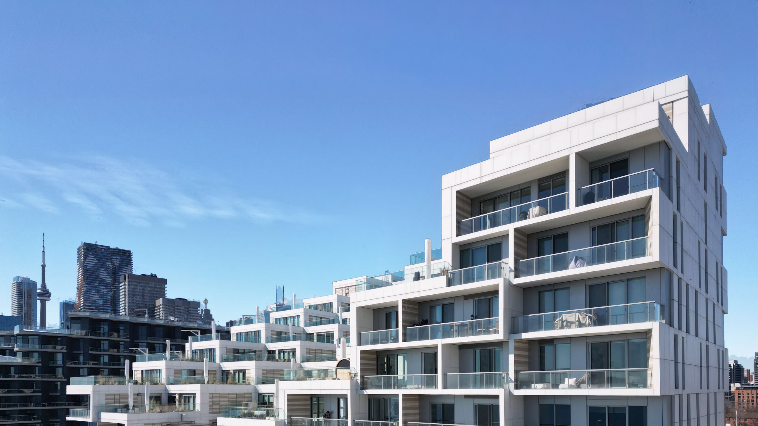 Modern stepped white apartment buildings with glass balconies under a clear blue sky, with a city skyline and a tall tower in the background.