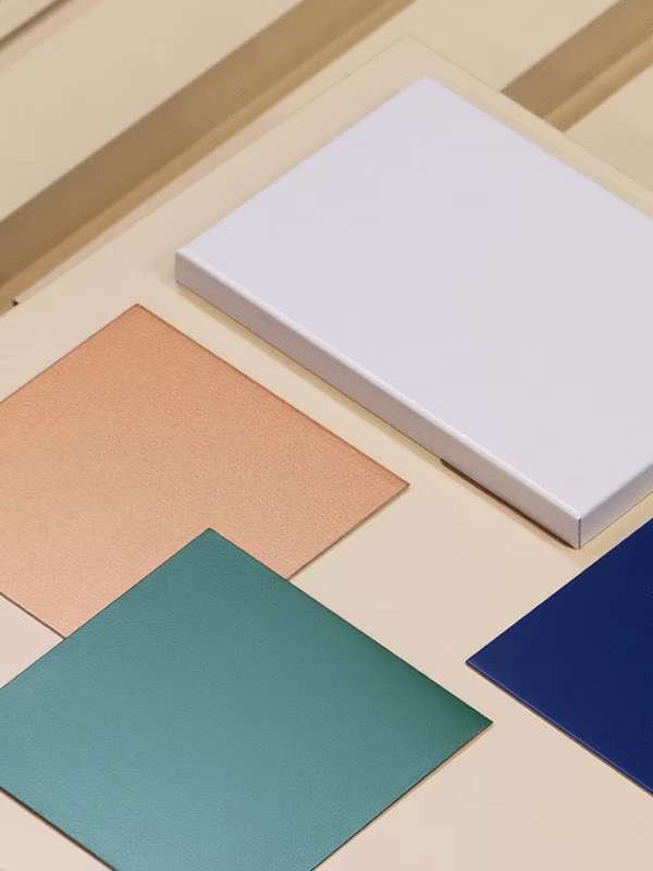Arrangement of four colored metal square sheets on a beige surface, displaying white, copper, teal, and dark blue colors.