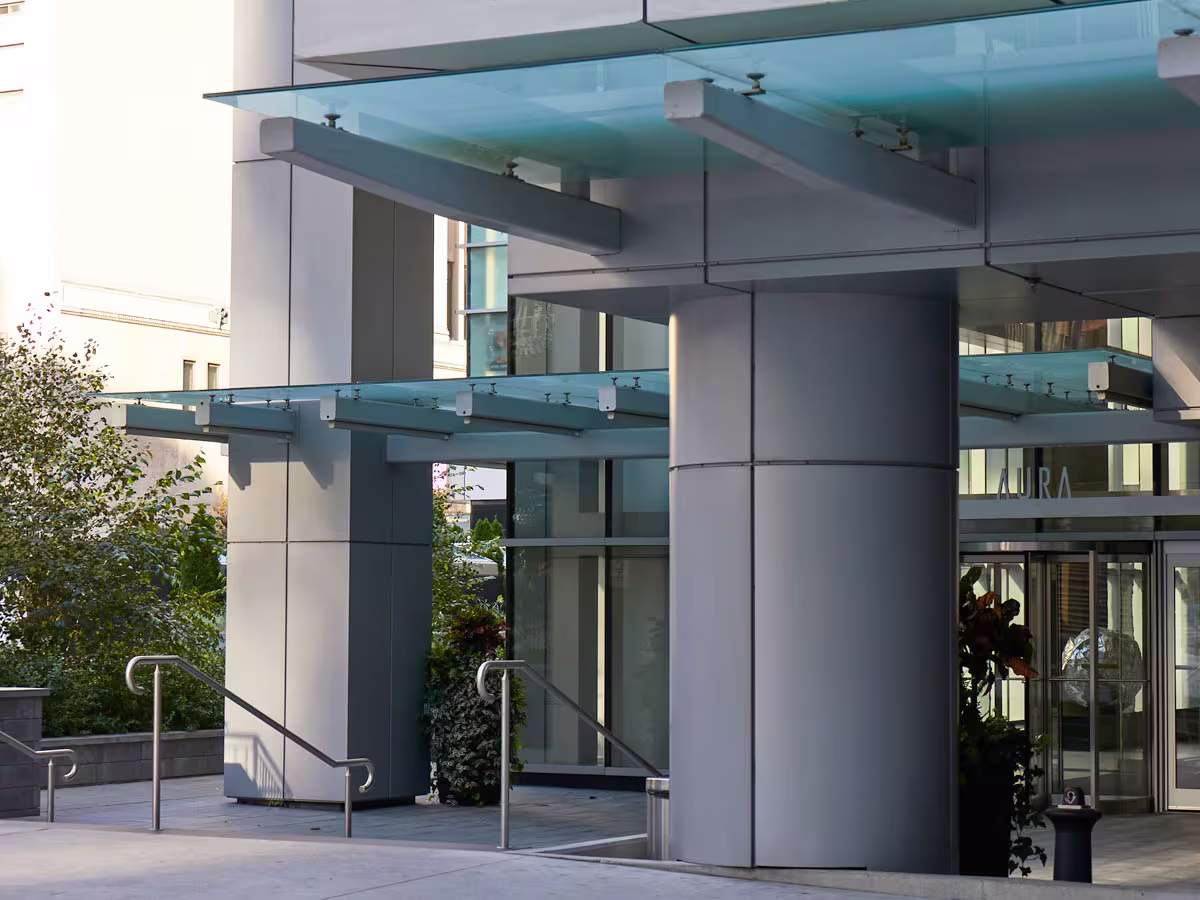 Modern building entrance with large cylindrical columns, glass doors, metal handrails along stairs, and a transparent glass canopy overhead, with greenery visible on the side.