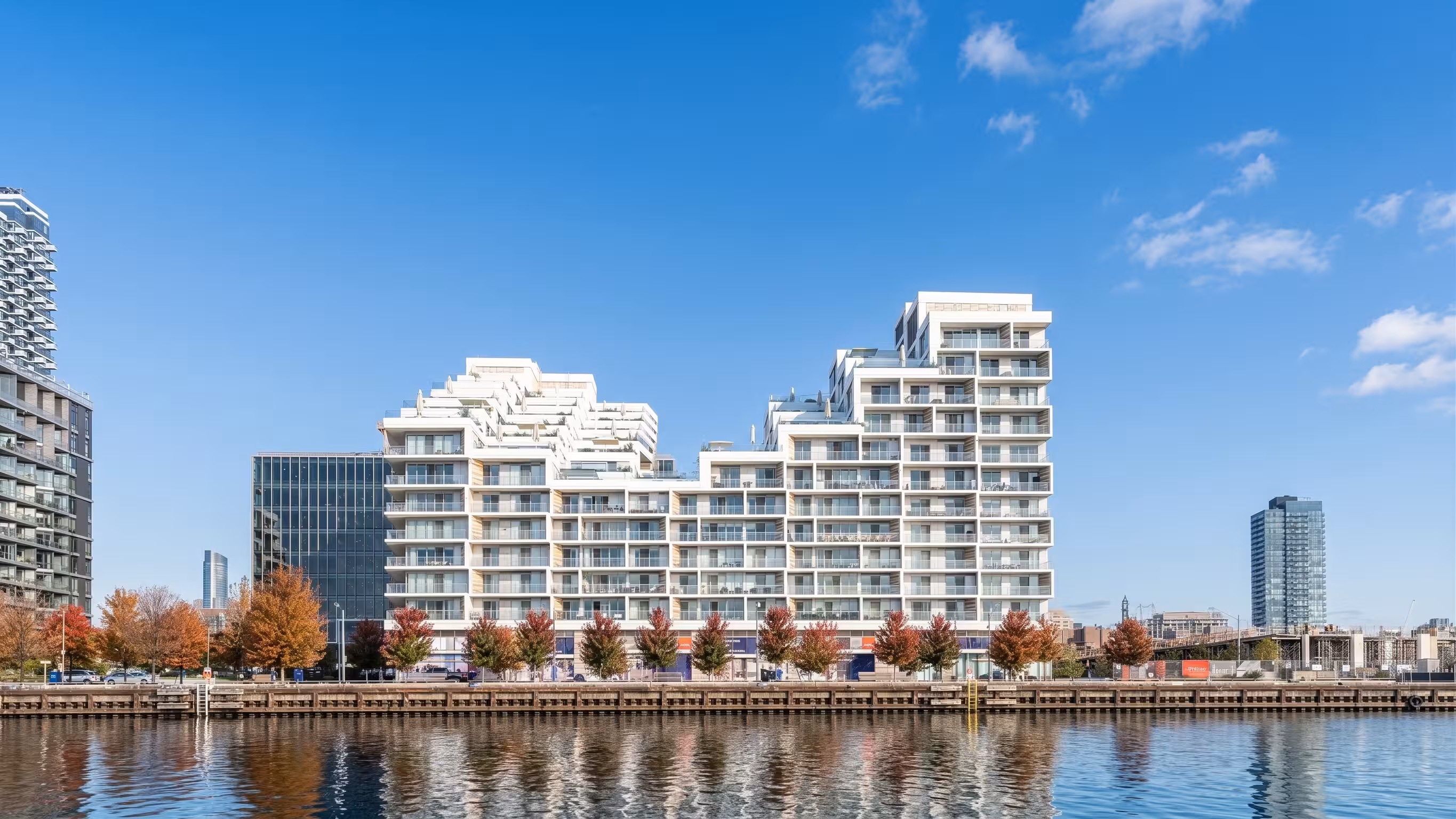 Modern white stepped apartment building beside waterfront with a clear blue sky and reflections in the water, surrounded by autumn trees and other high-rise buildings in the background.