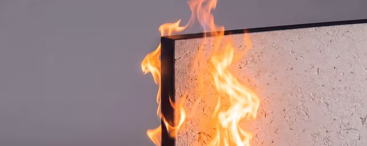 Flames rising along the corner edge of a textured wall panel with a gray background, illustrating a fire test.