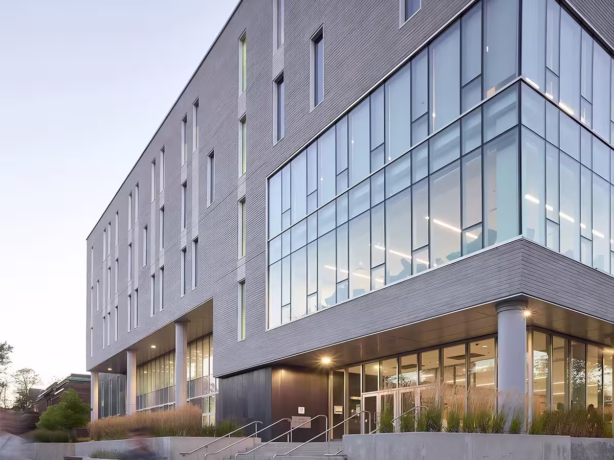 Modern commercial building with large glass windows on the corner and multiple floors, surrounded by greenery and outdoor lighting at dusk.
